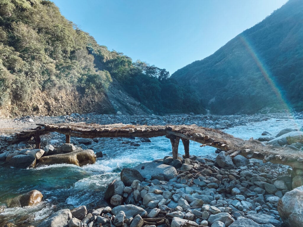 A wood and mud bridge over a river in Peru with mountains in the background