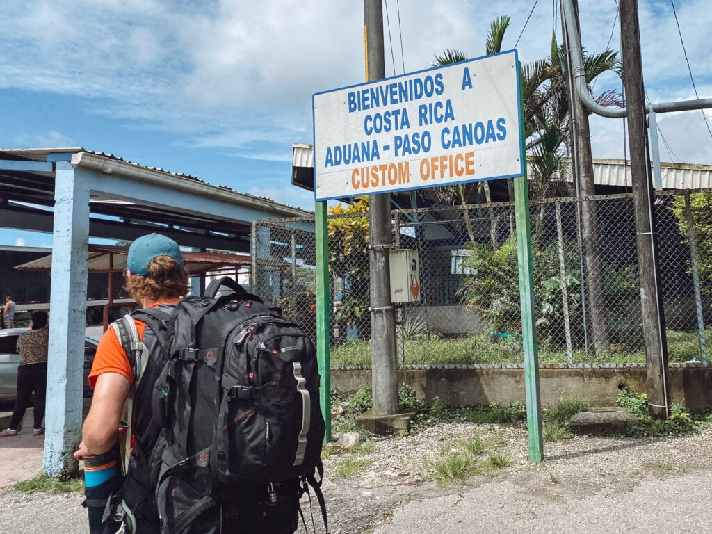 dan with big black backpack walking by a white blue and red sign that says bienvenidos a costa rica aduana - paso canoas custom office
