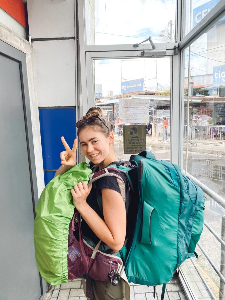 sarah giving the peace sign, wearing two big green backpacks on her front and back standing in the panama immigration office at the panama costa rica border
