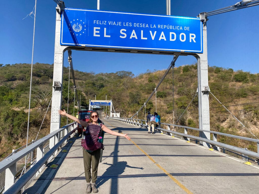 Sarah standing on a bridge under a blue sign that says feliz viaje les desea la republica de el salvador.