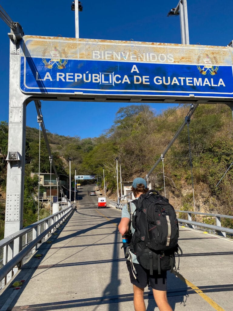 Dan wearing big backpacking crossing bridge under blue sign that says bienvenidos a la republica de guatemala