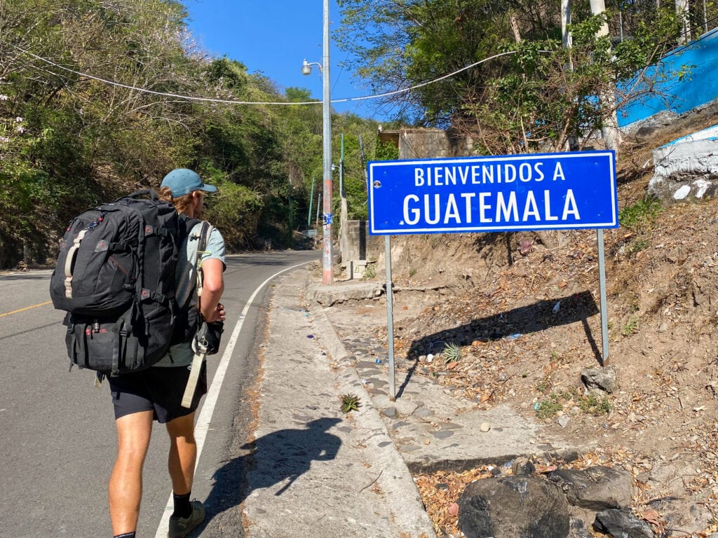 Dan walking with a big backpack on by a blue sign that says bienvenidos Guatemala at the Las Chinimas El Salvador to Guatemala border crossing