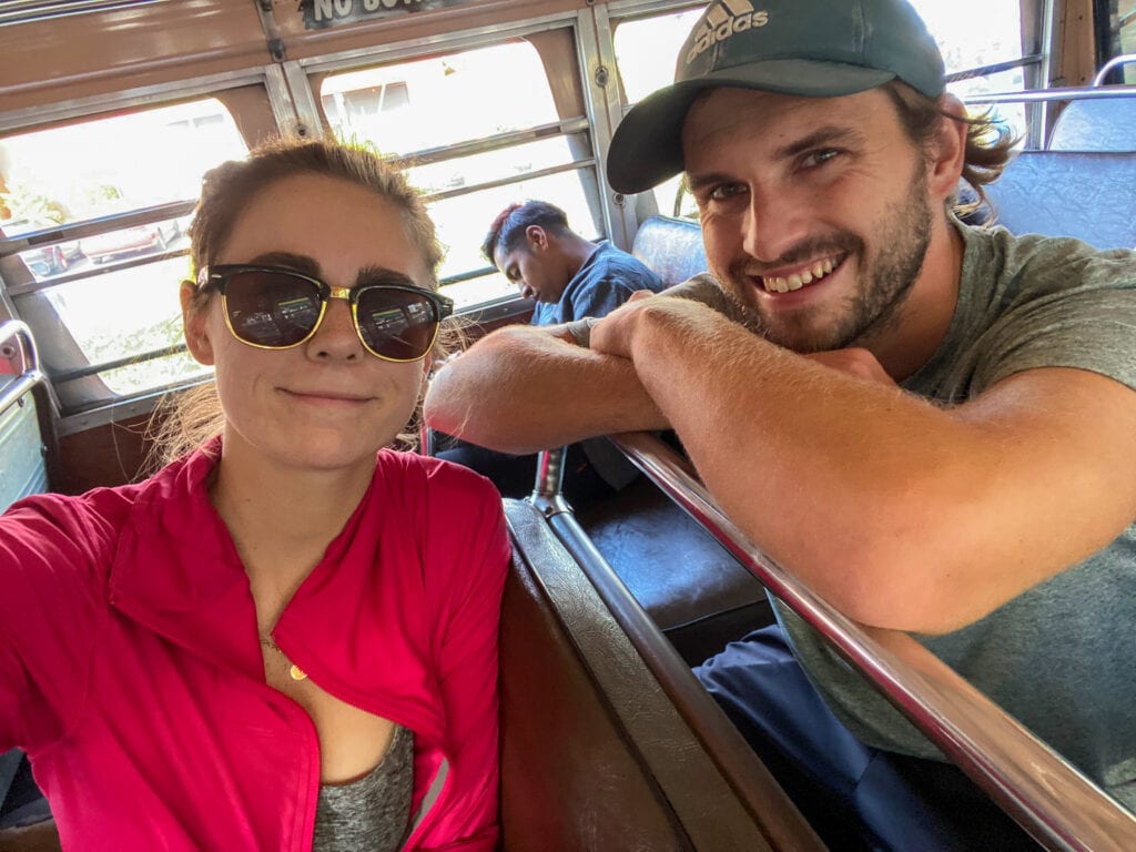 Dan and Sarah smile at the camera while leaning on the seats of a chicken bus. Sarah is wearing sunglasses and a pink jacket and Dan has a green hat and t-shirt on.