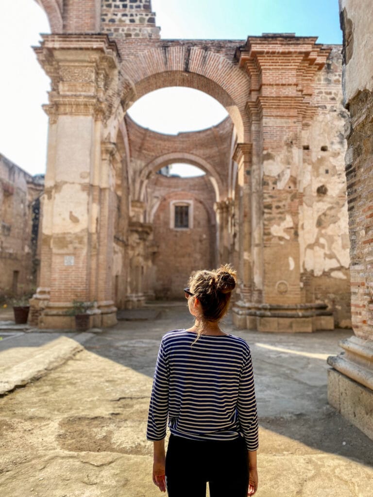 Sarah wears a striped shirt and faces away from the camera in side a large ruined cathedral.