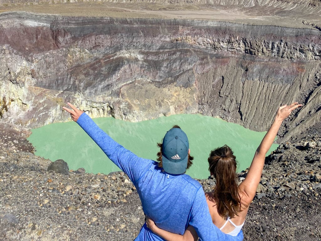 Dan (man in blue shirt and green hat) and Sarah (woman with brown hair in ponytail and white tank top) stand in front of blue green crater lake in volcano with their backs to the camera and hold their arms out with the peace sign.