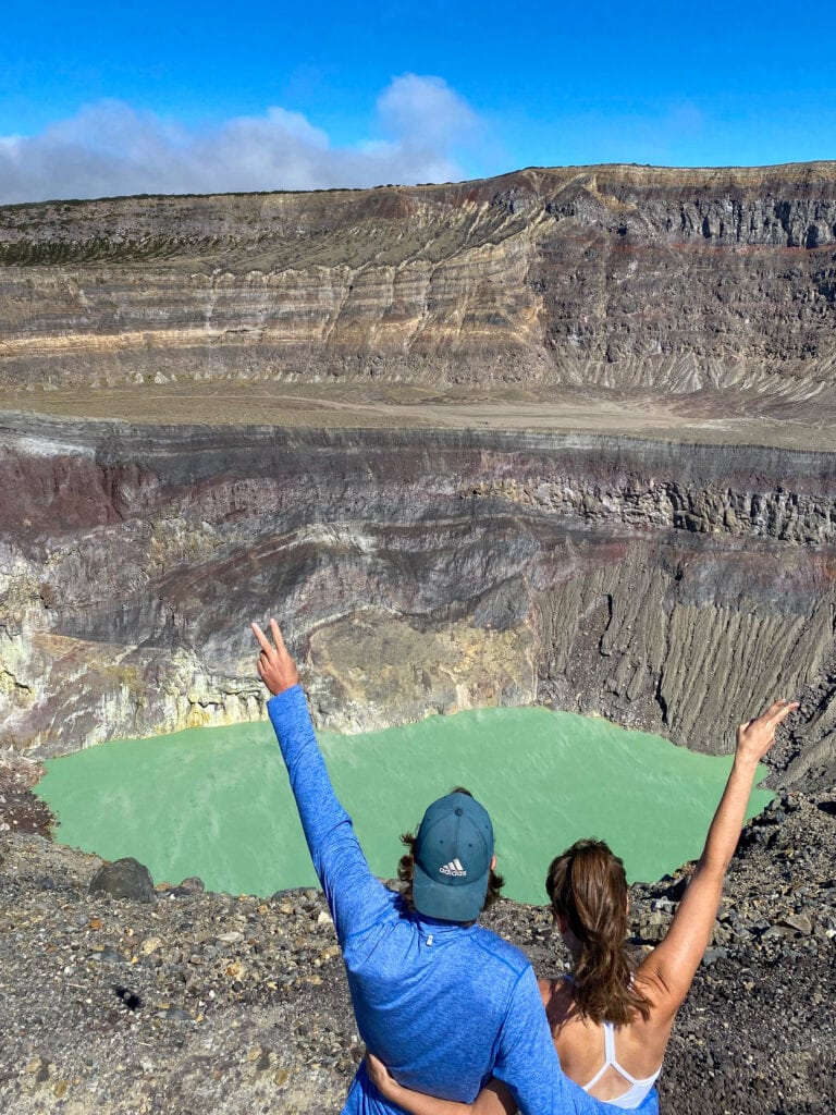 Dan (man in blue shirt and green hat) and Sarah (woman with brown hair in ponytail and white tank top) stand in front of blue green crater lake in volcano with their backs to the camera and hold their arms out with the peace sign.
