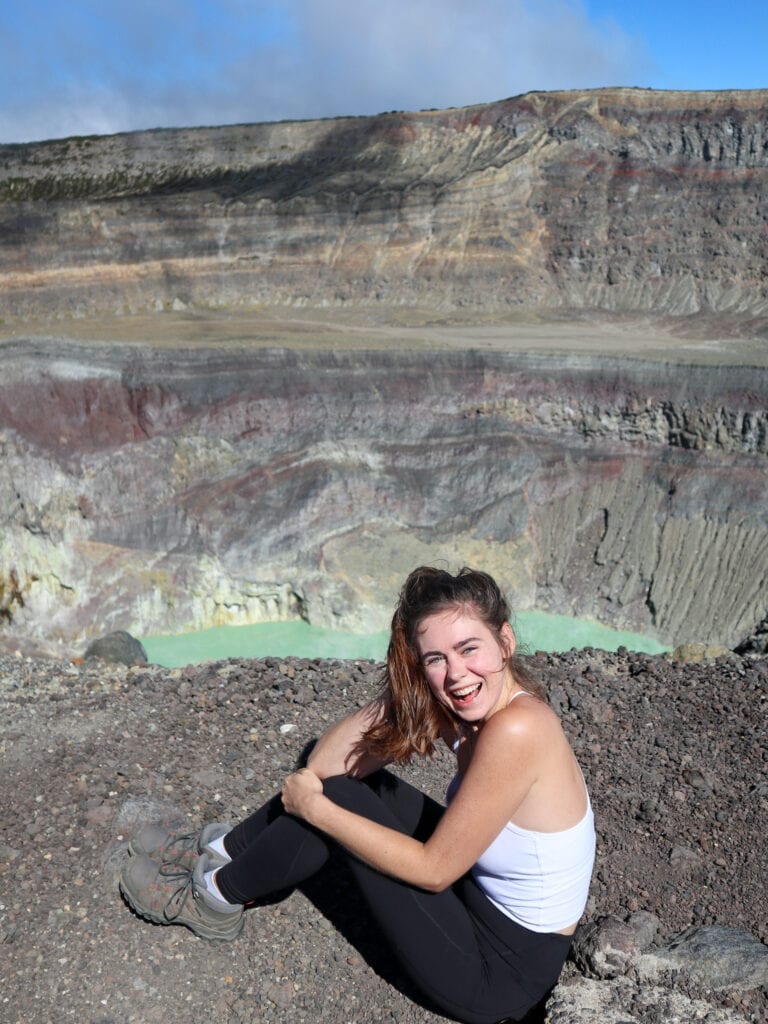 Sarah sits on ground and smiles for camera in front of Santa Ana Volcano crater lake.