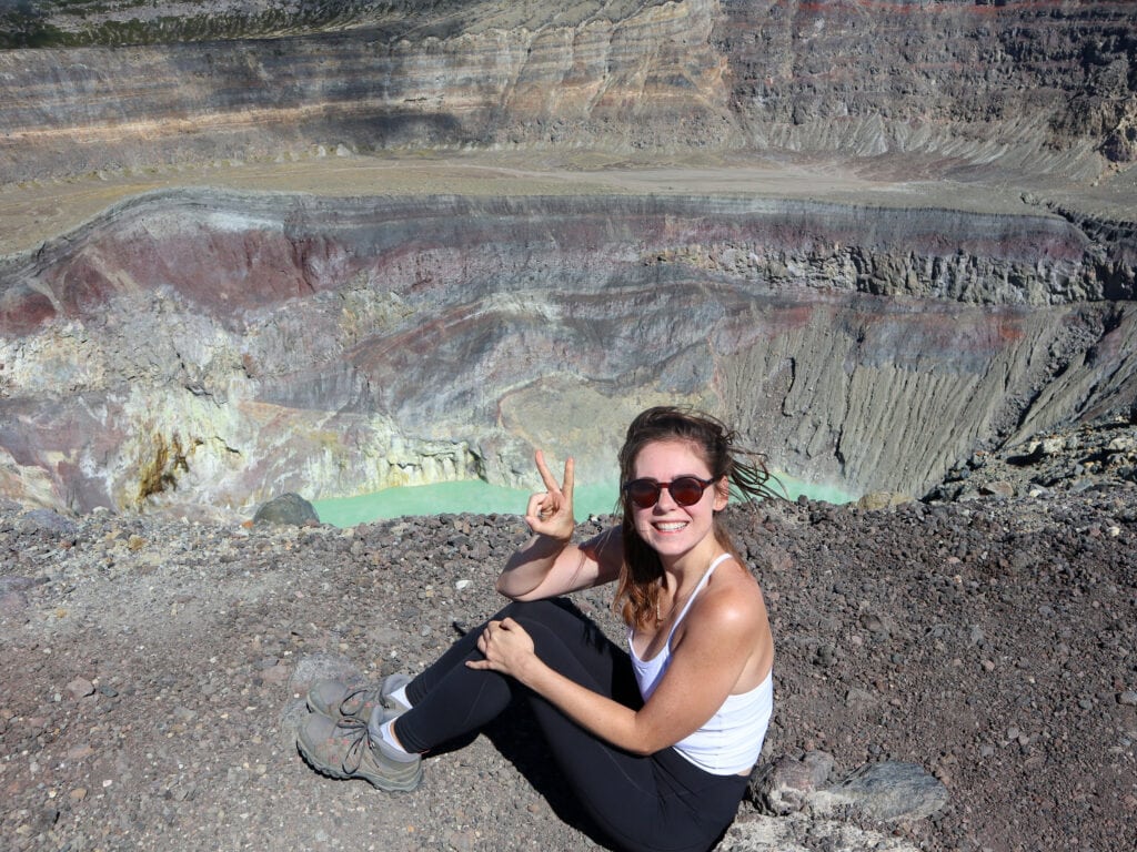 Sarah sits in front of the blue green crater lake of Santa Ana Volcano and smiles and holds up a peace sign