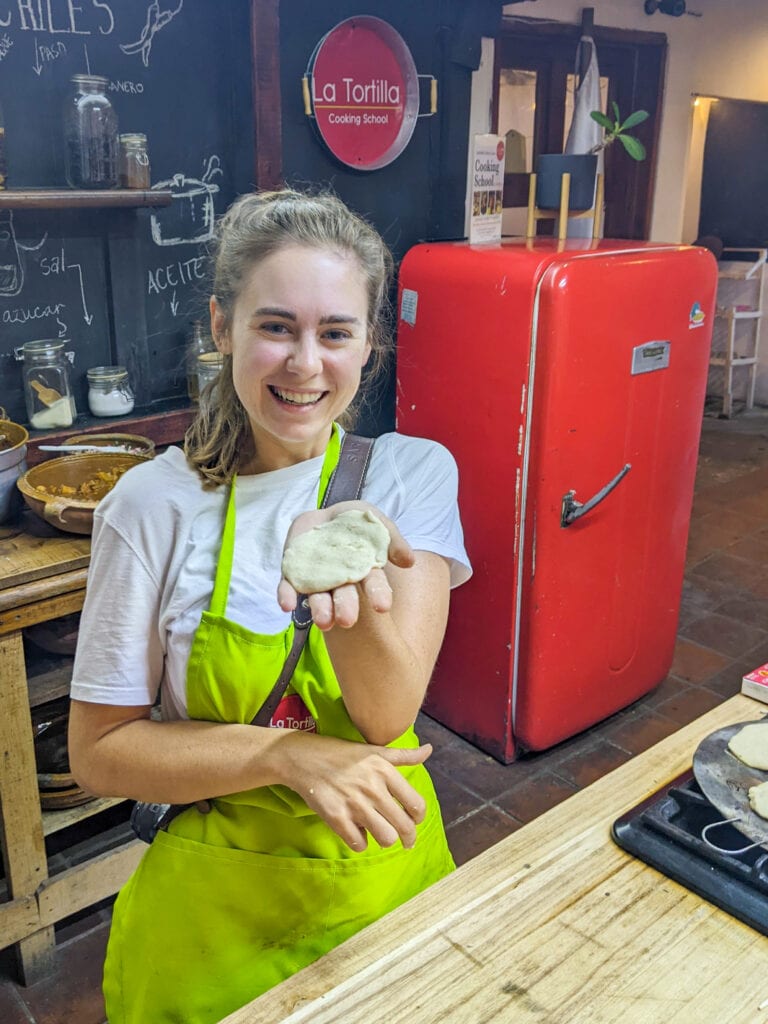 Sarah wears a green apron, smiles at the camera, and holds an uncooked tortilla dough in her palm.