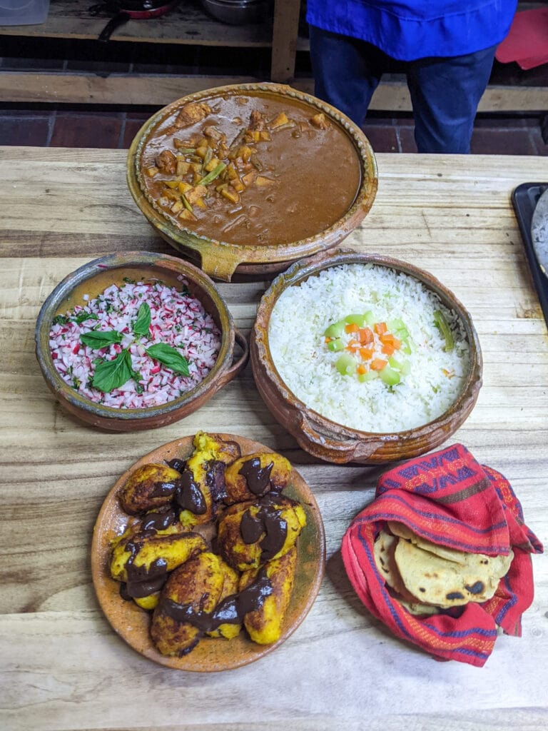 An overhead shot of five clay bowls with pepian, radish salad, rice, rellenitos, and tortillas.