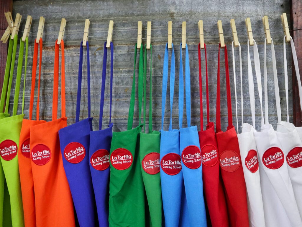 A row of colorful aprons with red circular badges that say La Tortilla Cooking School on them.