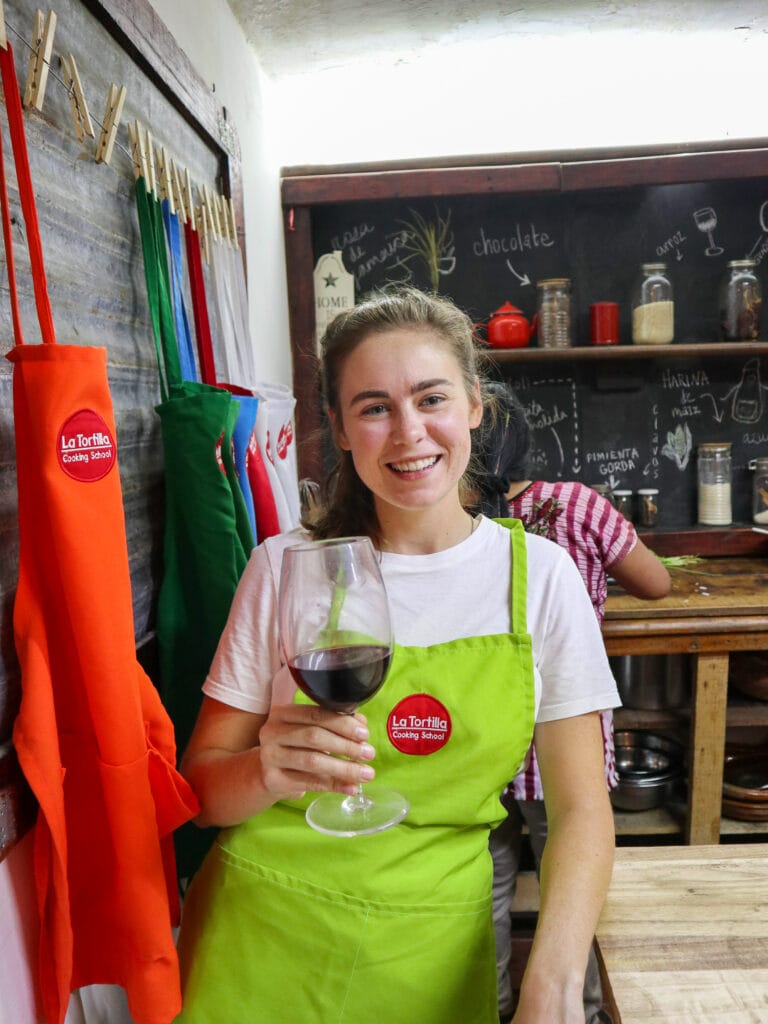 Sarah wears a green apron, holds a glass of red wine, and smiles at the camera.