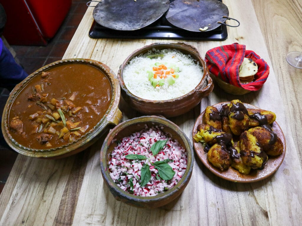 An overhead shot of five clay bowls with pepian, radish salad, rice, rellenitos, and tortillas.