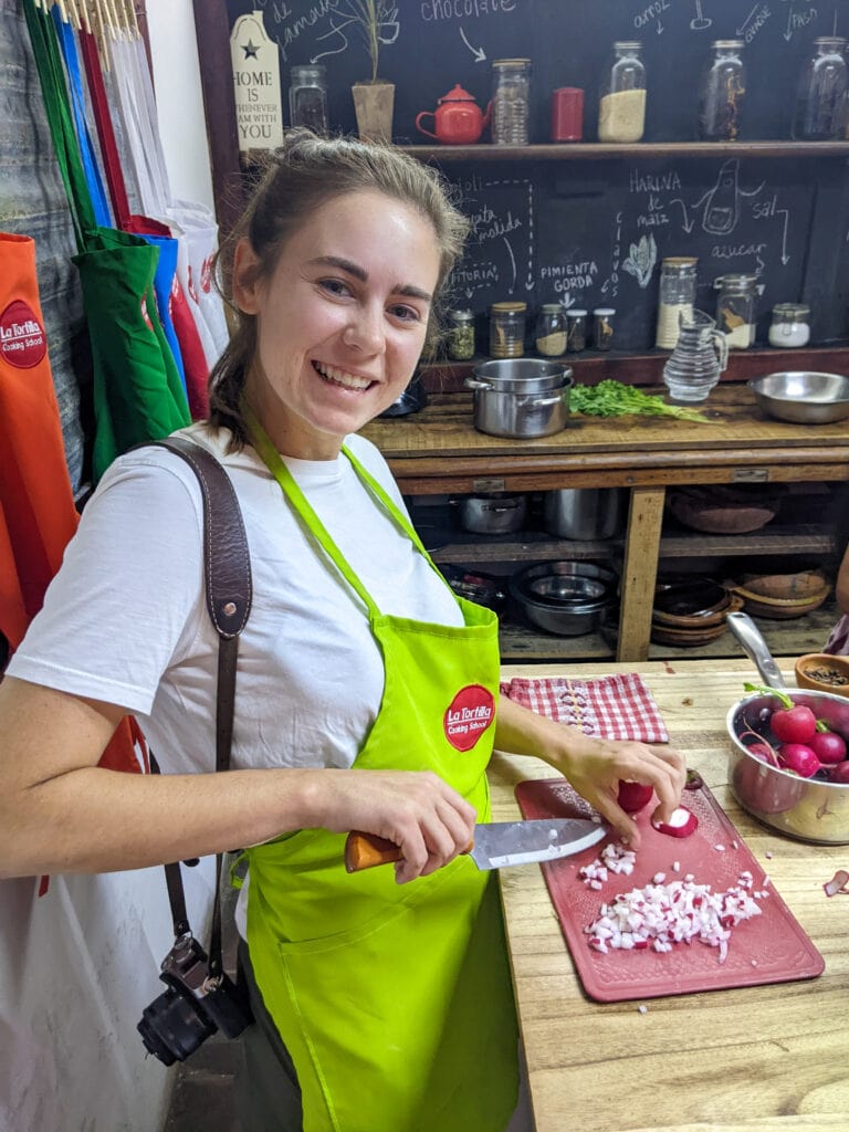 Sarah wears a green apron, cuts radishes on a cutting board, and smiles at the camera.