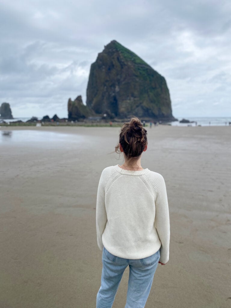 Sarah wearing white sweater and jeans facing away from the camera standing in front of Haystack Rock