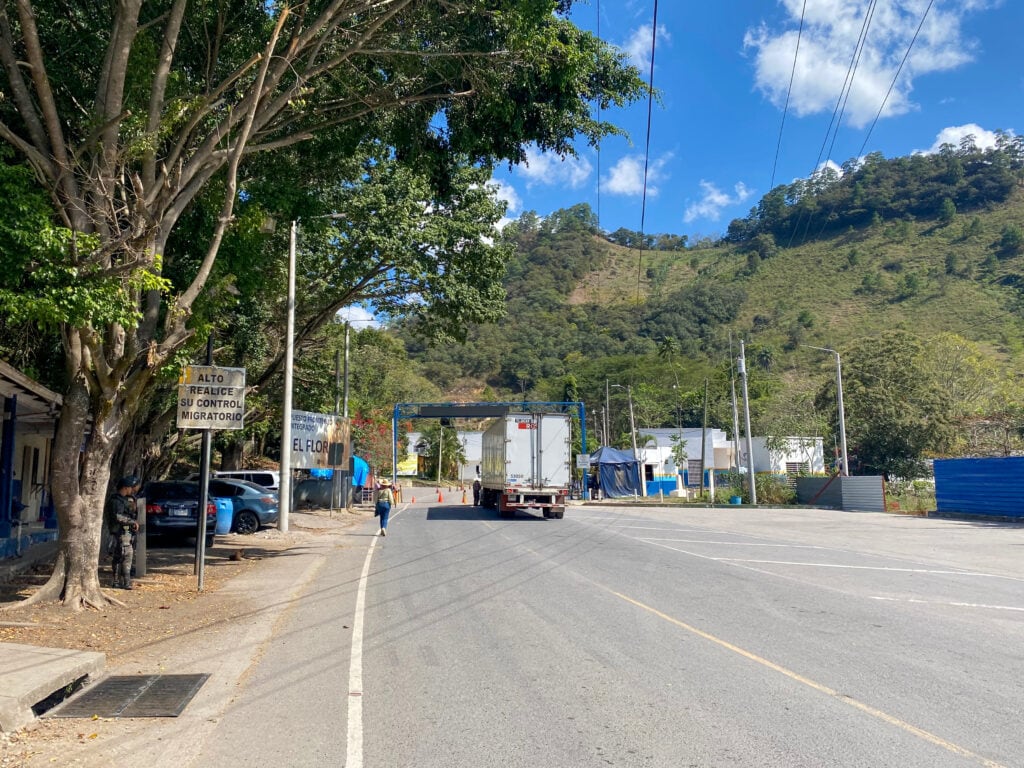 A paved road with green hills in the back and a semi truck. In the distance is a blue overhead gate and a sign that says "el florido".
