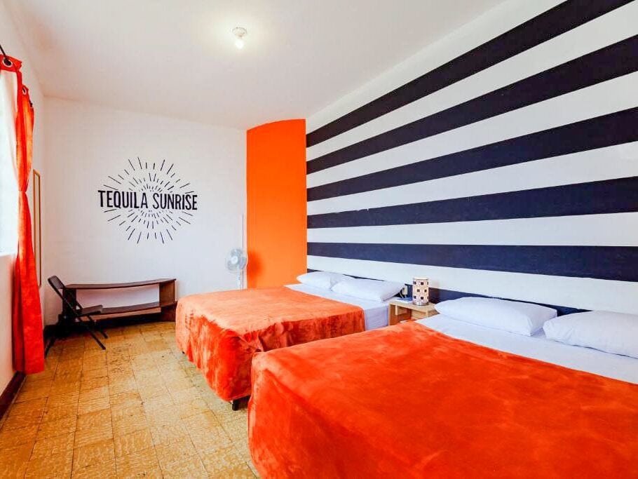 A hostel room with two orange beds. There is a sign on the far wall that says Tequila sunrise.