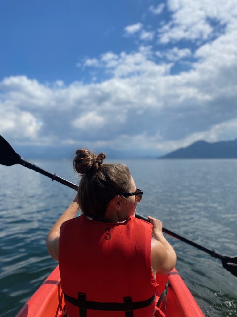 The back of Sarah's head as she paddles in a kayak in a large lake. She has on sunglasses and an orange life vest.