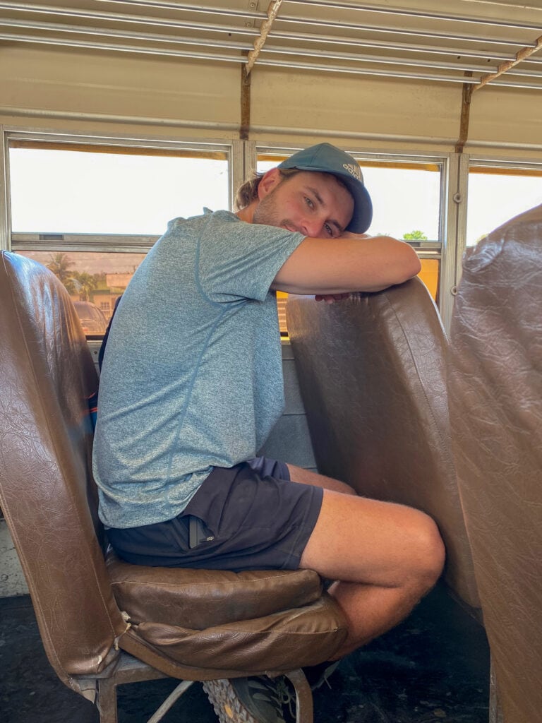 Dan, a white man in a green shirt and green hat, sits on a brown bus seat and leans on the seat in front of him and looks toward the camera.