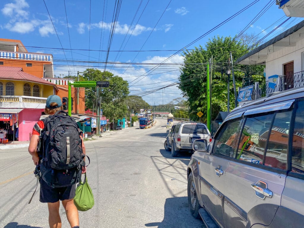 A sunny street with a bridge in the distance. Dan is walking away from the camera, wearing a large black backpack and carrying a green bag.