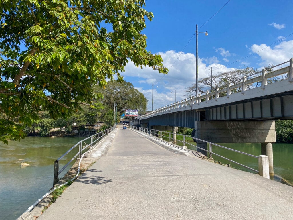 An asphalt bridge over a river, with another bridge visible above and to the right.