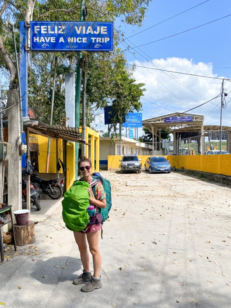 Sarah standing under a sign that says "feliz viaje have a nice trip" at the guatemala belize border.