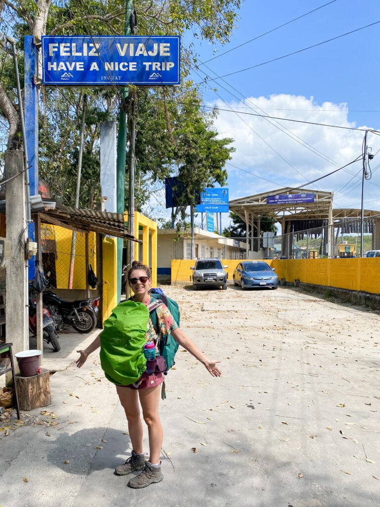 Sarah standing under a sign that says "feliz viaje have a nice trip" at the guatemala belize border.