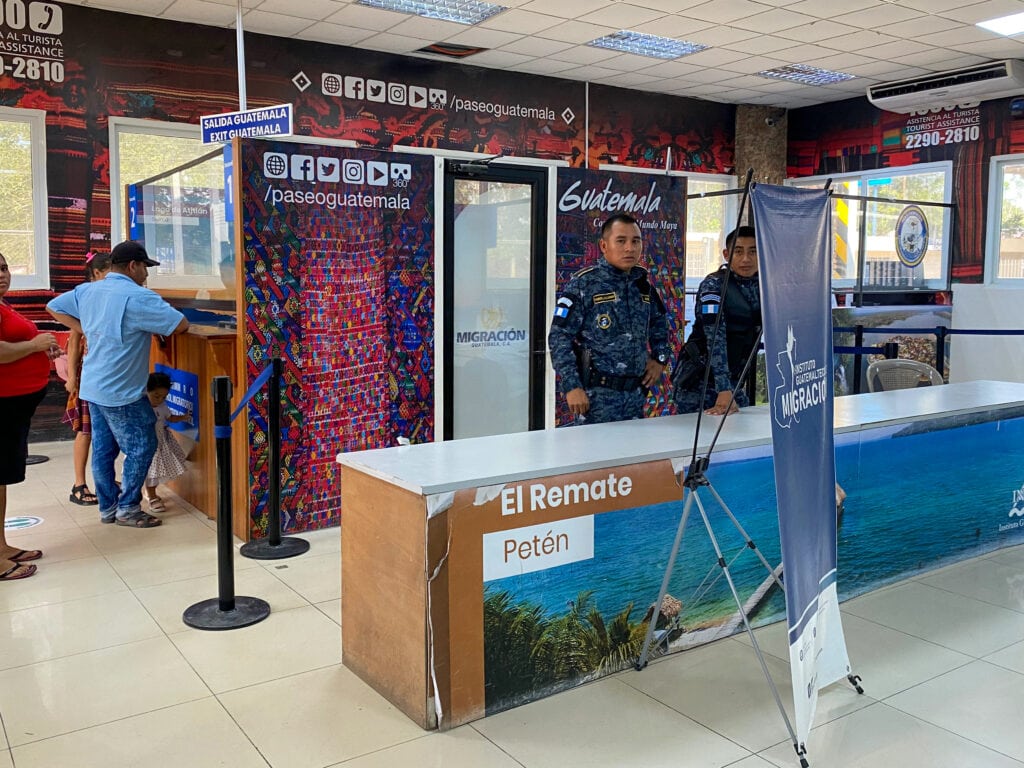 Guatemala immigration building. Two men in camo holding rifles with Guatemalan flags on their upper arms look at the camera.