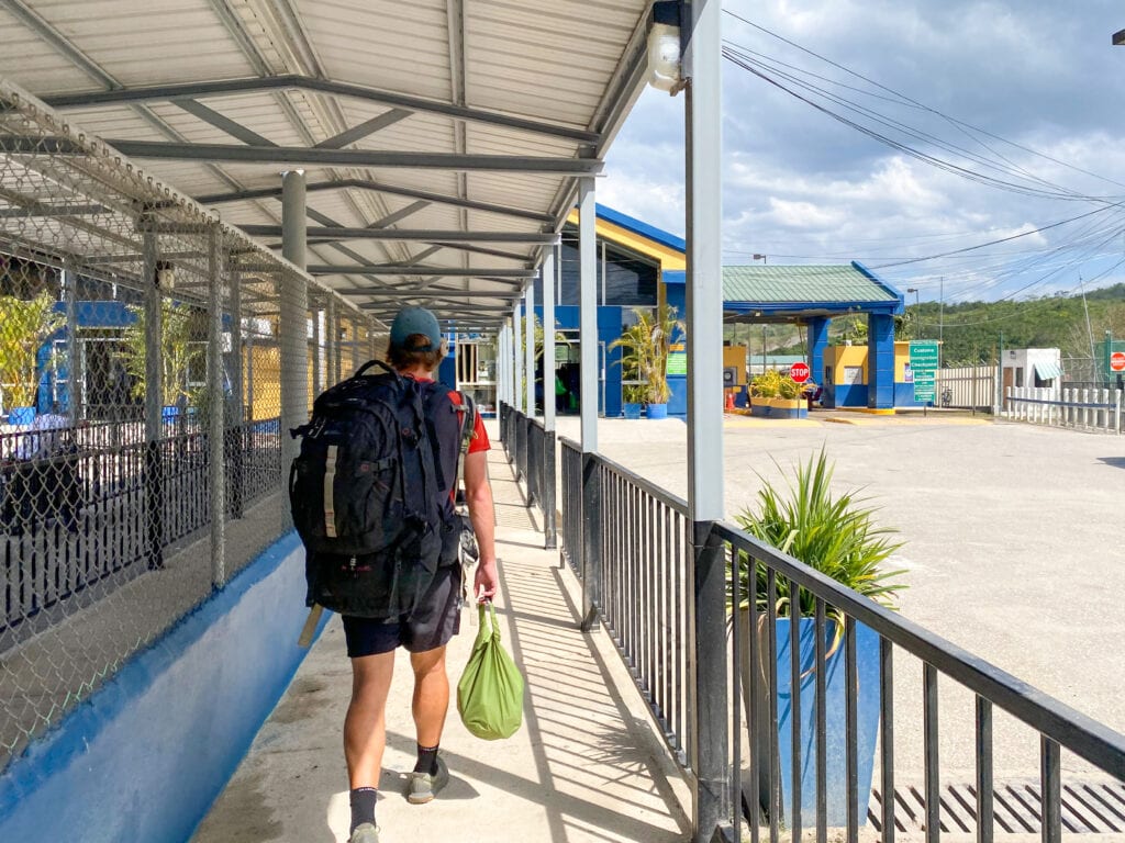 A shaded walkway with a railing to the right-hand side, with a blue and yellow building in the distance. Dan walks with his back to the camera, wearing a big black backpack.