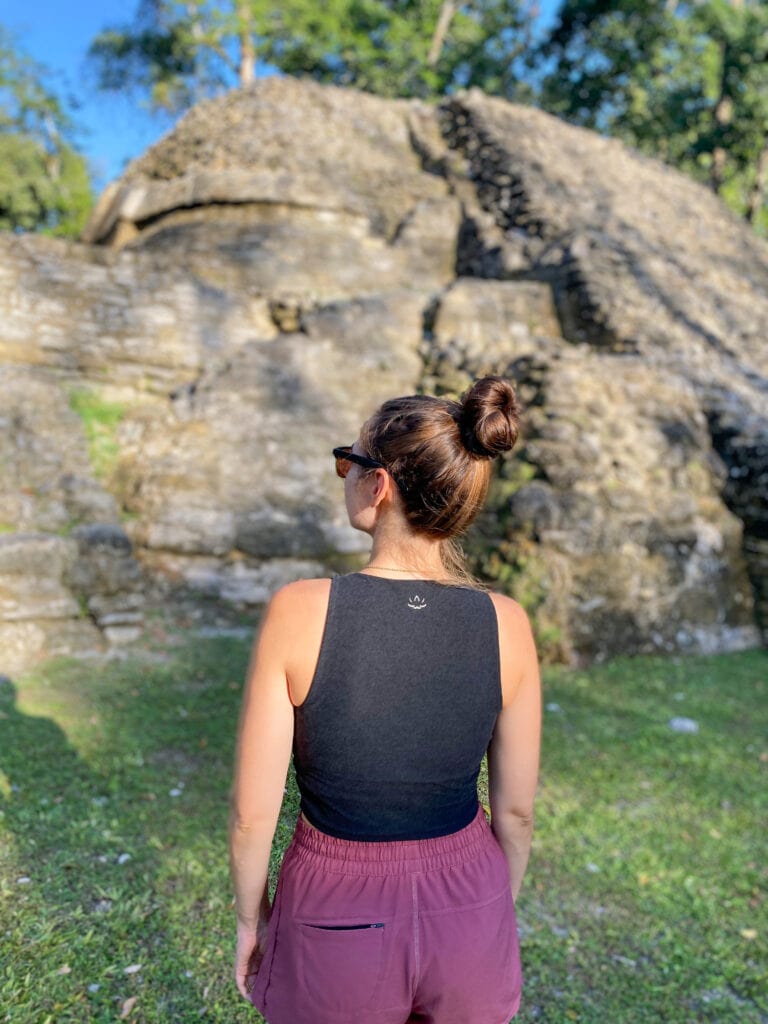 Sarah stands looking away from the camera and toward the ruins at Cahal Pech. Her hair is in a bun and she wears sunglasses, a black tank top, and dark pink shorts.