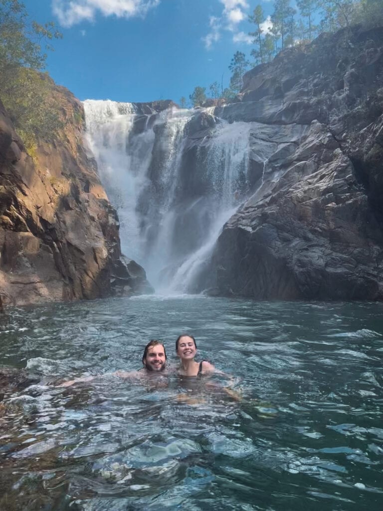 Sarah and Dan smiling and swimming in a natural pool in front of a large waterfall.