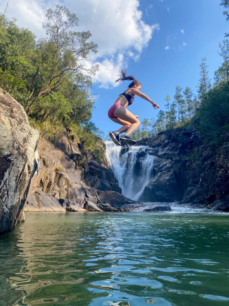 Sarah jumps off a cliff into a natural pool with Big Rock waterfall in the background.