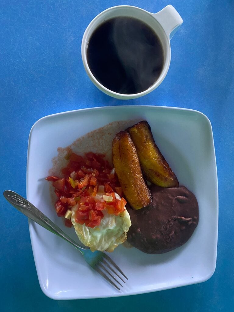 An overhead shot of a breakfast of hot steaming coffee and a plate of plantains, red salsa, refried beans, and egg.