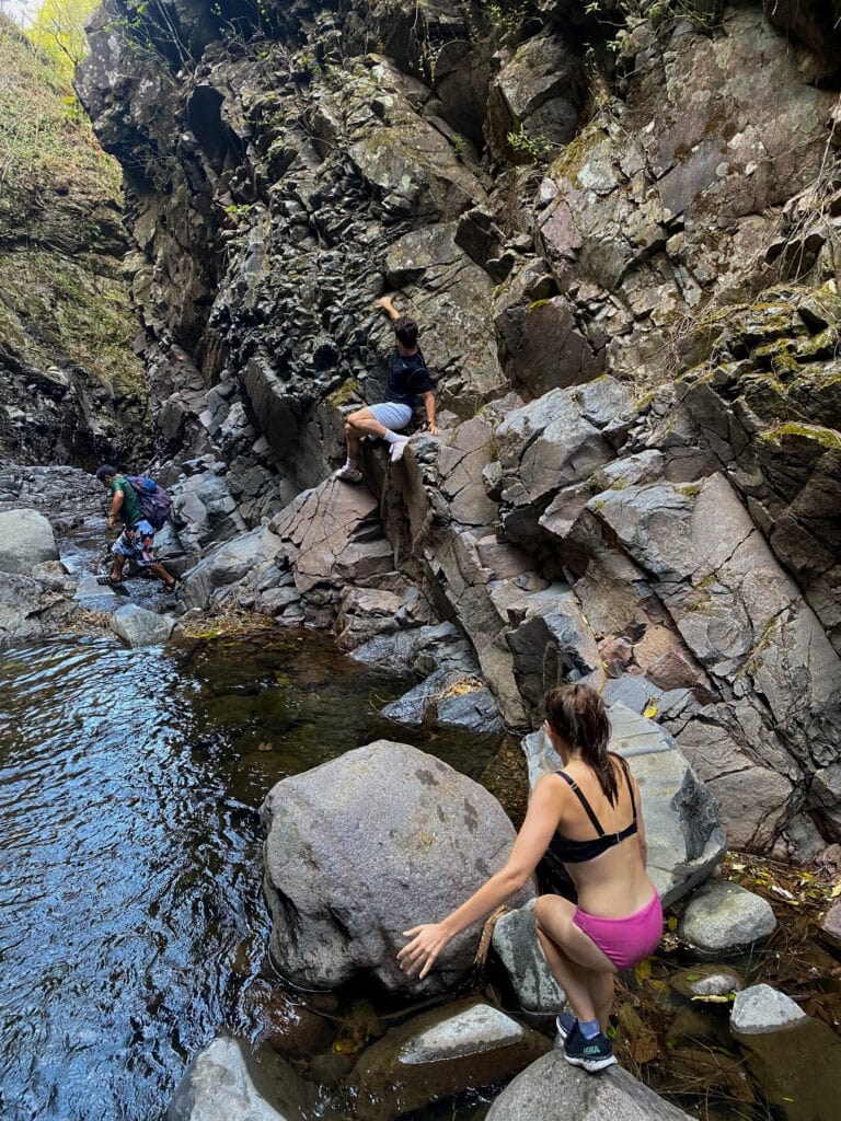 Sarah is in the foreground climbing between two small boulders and in the distance another traveler is with his back flat against the rock wall, and William the tour guide walks across a river.