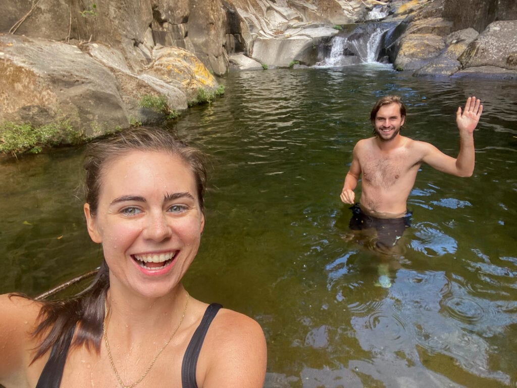 A selfie of Sarah and Dan in a river. Sarah smiles and Dan is in the back waving.