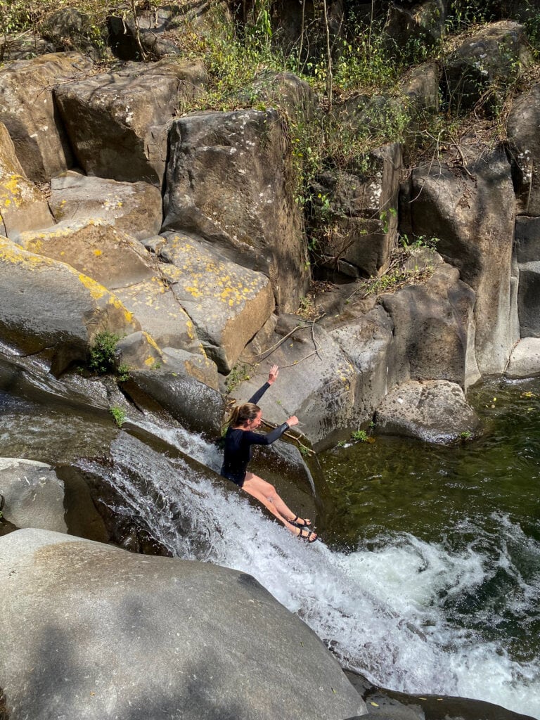 A girl in a black swimsuit slides down a natural slide in El Salvador with her arms up in the air.