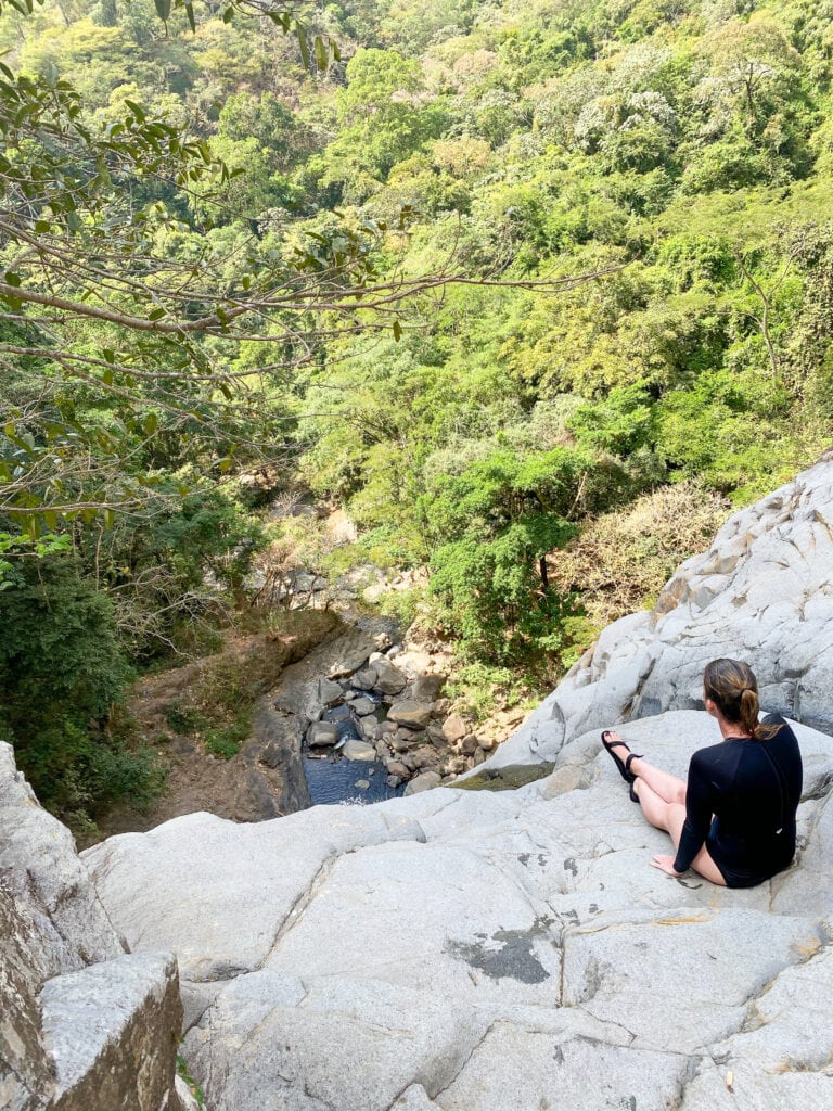 A girl in a black shirt and bikini bottoms and sandals sits at the top of the waterfall looking out.