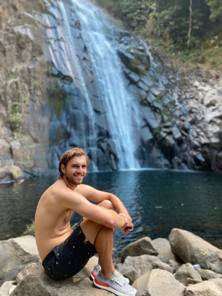 Dan sits in front of the waterfall and smiles at the camera.