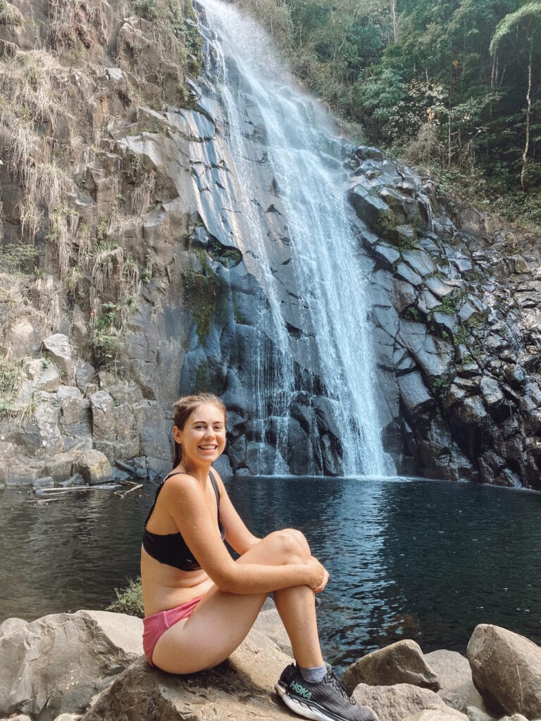Sarah sits on a boulder by a waterfall, wearing a bikini and black sneakers, and smiles at the camera.