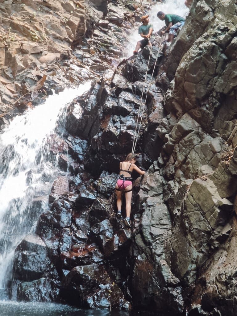 Sarah in a harness rock climbing down a cliff face next to a waterfall. You can see William and Edwin holding the ropes at the top.