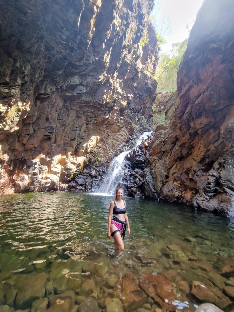 Sarah stands in the water and smiles at the camera, Dan climbs down the waterfall in the distance.