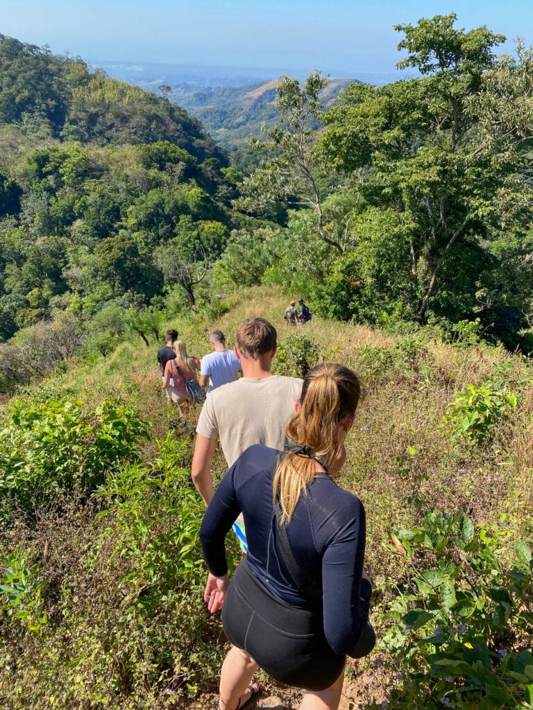 A group of travelers hikes downhill through a grassy field with forest in the distance in El Impossible National Park in El Salvador.
