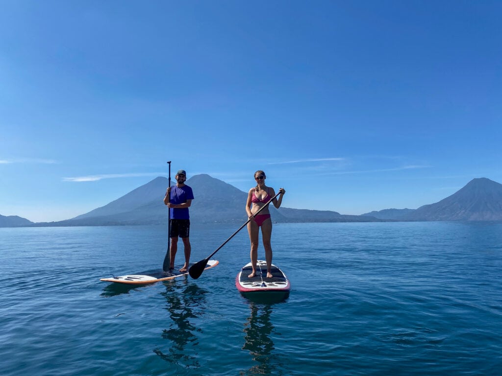 Sarah and Dan stand on paddle boards in Lake Atitlan and smile at the camera.