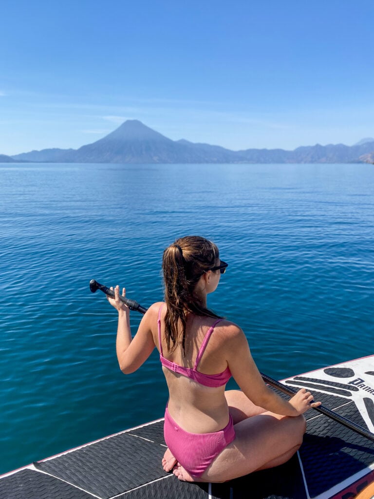 Sarah in pink bikini sits on SUP facing away from camera in Lake Atitlan with volcano in the background.