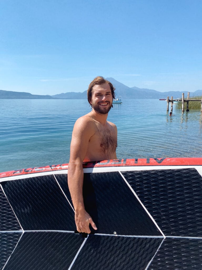 Dan smiles at camera holding a SUP on beach by Lake Atitlan.