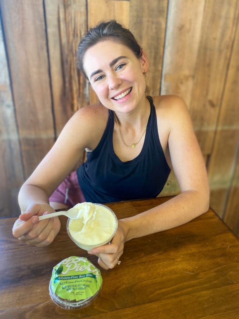 Sarah, a white woman with brown hair and a black tank top, holds the gluten free key lime pie in a cup and smiles at the camera.
