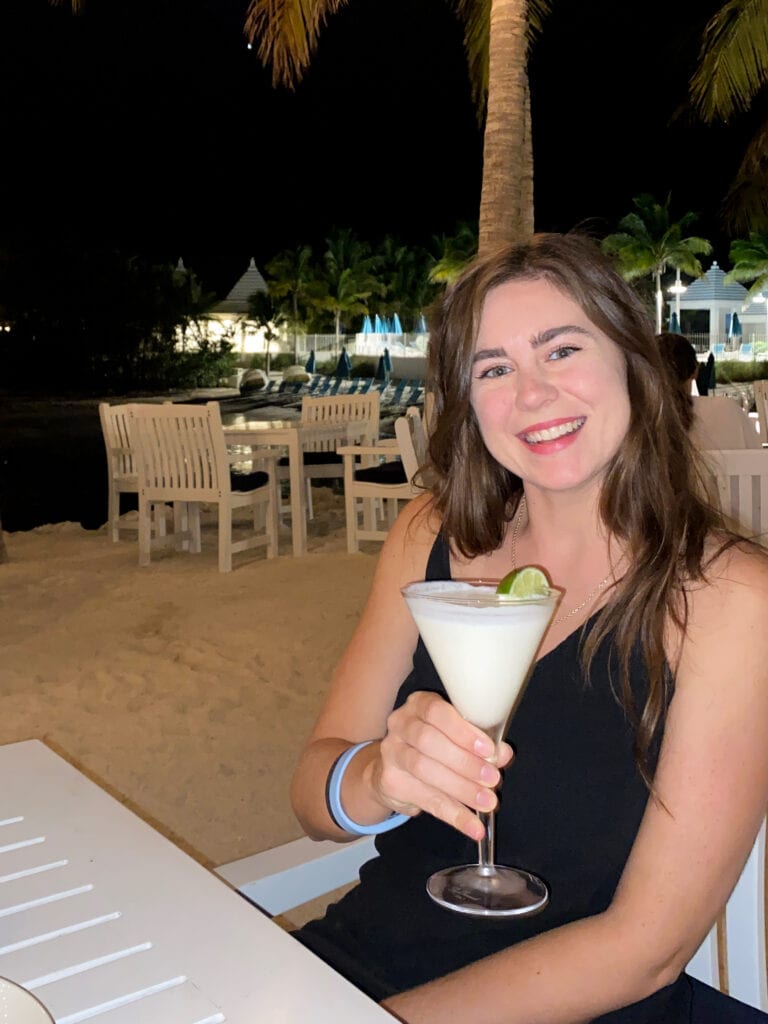 Sarah, a white woman with brown hair wearing a black dress, holds a light green key lime martini and smiles at the camera.