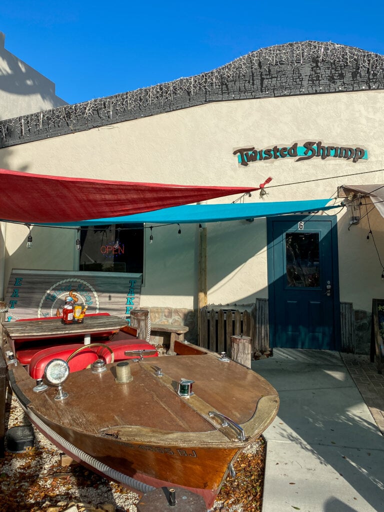 The exterior of Twisted Shrimp restaurant. A sign reads 'twisted shrimp' and there is a wooden boat reconverted into a red picnic table.