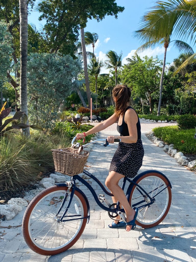 Sarah wears a black tank top and black and white skirt and sits on a bike with a wicker basket, surrounded by palm trees.