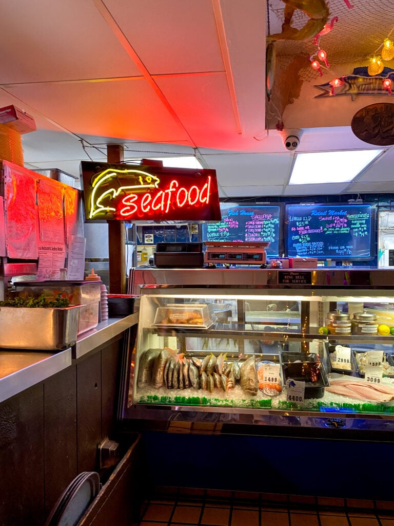 A red neon sign that says 'seafood' behind a bar with a glass tank of fresh whole fish.